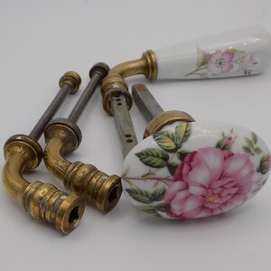 Könnte beinhalten: Satz von vier antiken Türklinken mit Porzellan-Blumendesigns. Die Knöpfe haben einen weißen Hintergrund mit rosa und grünen Rosen. Die Knöpfe sind aus Messing und haben eine polierte Oberfläche.