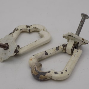 Puede incluir: Dos tiradores de cajón de metal blanco con una abertura en forma de corazón. Los tiradores tienen un acabado desgastado y están unidos a tornillos.