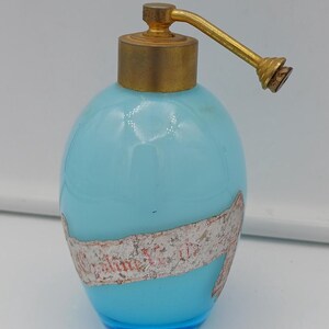 Peut inclure: Un flacon de parfum bleu clair vintage avec un atomiseur et un bouchon dorés. La bouteille a une étiquette enroulée autour. La bouteille est de forme ovale et repose sur une surface blanche.