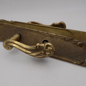 Puede incluir: Una manija de puerta de latón con un diseño decorativo y ornamentado. La manija tiene una forma curva y una cerradura.