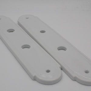 Puede incluir: Dos placas de puerta rectangulares blancas con bordes redondeados y cuatro agujeros cada una. Las placas tienen un diseño decorativo alrededor de los bordes.