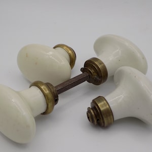 Può includere: Tre pomelli per porte in porcellana bianca con dettagli in ottone e un perno in metallo. I pomelli sono di forma ovale e hanno una finitura liscia.