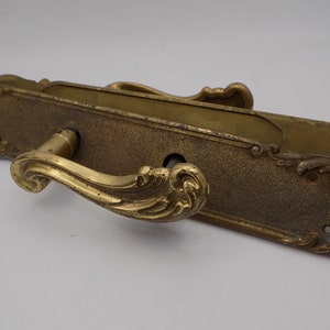 Può includere: Una maniglia per porta in ottone con un design decorativo e ornato. La maniglia ha una forma curva e una serratura. La piastra di fondo è rettangolare con un design ornato simile.