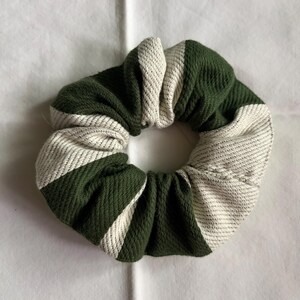 Puede incluir: Una scrunchie a rayas verdes y blancas. El accesorio para el cabello está hecho de una tela texturizada y presenta rayas diagonales alternas de color verde oscuro y blanco roto. La scrunchie es circular y parece nueva.