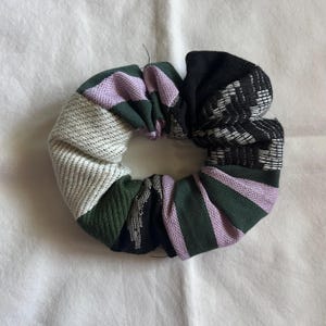 Puede incluir: Una scrunchie hecha a mano con un diseño de patchwork. La tela es una mezcla de colores, incluyendo negro, verde, lavanda y blanco. La scrunchie está hecha de varias telas estampadas, creando un aspecto único y texturizado. Este accesorio para el cabello es perfecto.