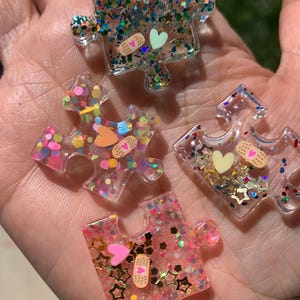 Könnte beinhalten: Vier Puzzleteil-Anhänger aus klarem Kunstharz mit eingebettetem buntem Glitzer, kleinen sternförmigen Pailletten, Miniaturherzen und Pflastermotiven mit rosa Herzen. Jedes Teil hat eine glänzende Oberfläche und individuelle Dekorationen.