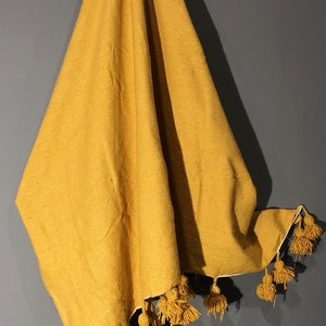 May include: A mustard yellow throw blanket with a fringe of tassels. The blanket is folded and hanging on a wall.