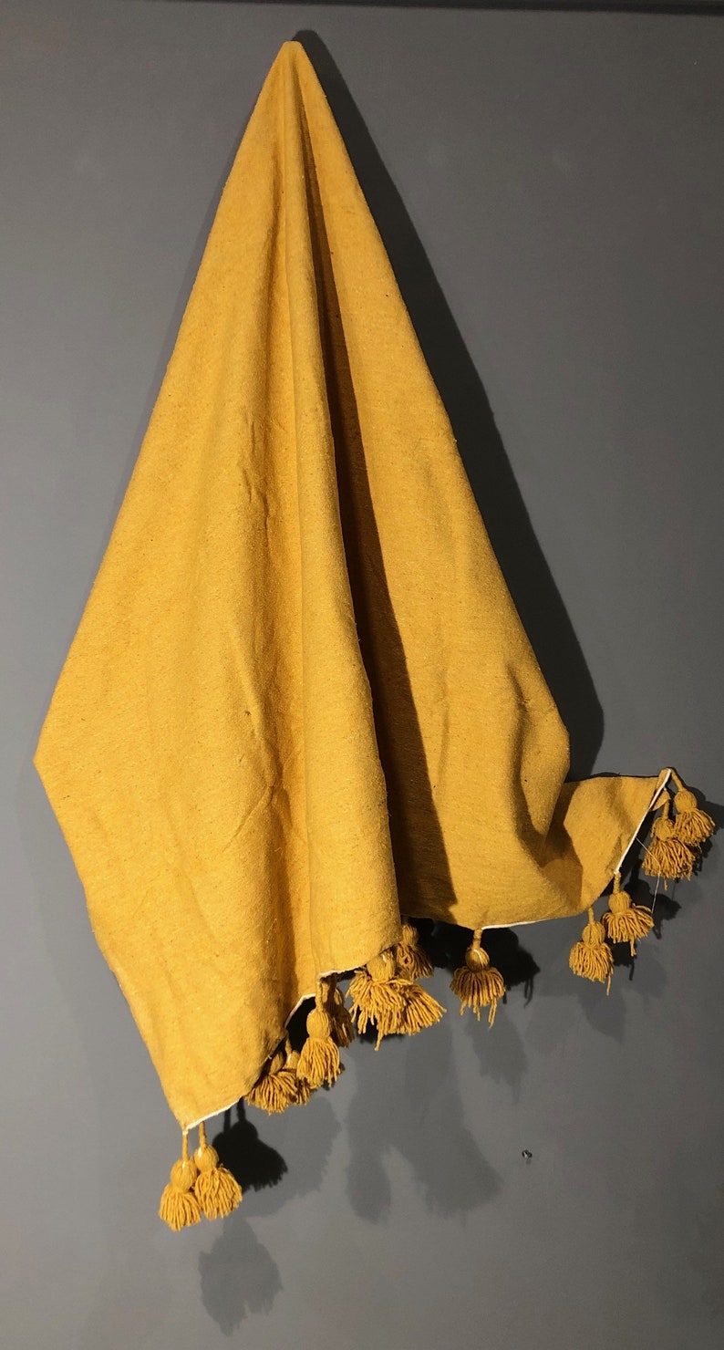 May include: A mustard yellow throw blanket with a fringe of tassels. The blanket is folded and hanging on a wall.