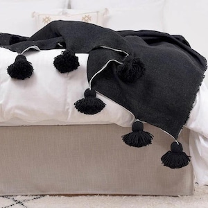 May include: Black throw blanket with white trim and black pom-poms. The blanket is draped over a bed with a white bedspread and a beige bed skirt.