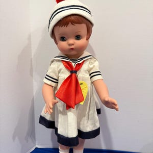 Könnte beinhalten: Eine Vintage-Puppe in einem Matrosen-Outfit. Die Puppe hat rotes Haar und blaue Augen und trägt eine weiße Matrosenmütze mit marineblauen Streifen und einem roten Bommel. Das Outfit umfasst ein weißes Kleid mit marineblauen Verzierungen, eine rote Krawatte und rote Schuhe.