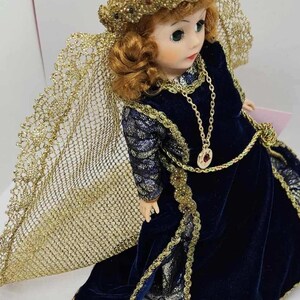 May include: A porcelain doll with auburn hair, green eyes, and a serene expression. The doll wears a dark blue velvet gown with gold trim and a matching gold necklace. A gold-colored veil and crown complete the elegant ensemble.
