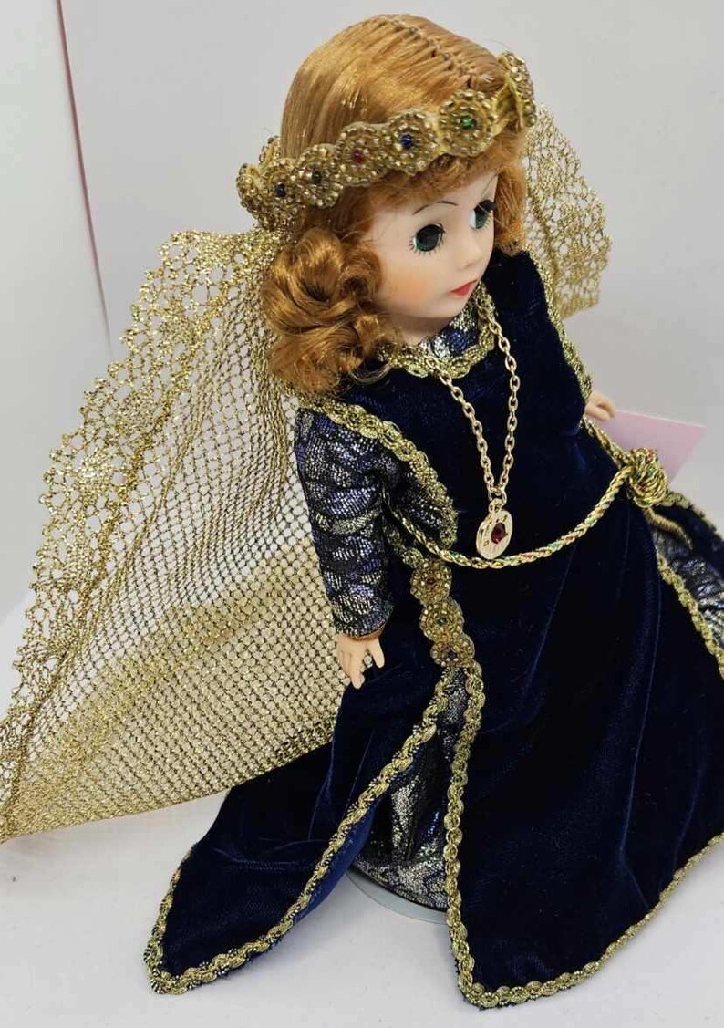 May include: A porcelain doll with auburn hair, green eyes, and a serene expression. The doll wears a dark blue velvet gown with gold trim and a matching gold necklace. A gold-colored veil and crown complete the elegant ensemble.