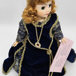May include: A vintage doll with auburn hair and green eyes, wearing a dark blue velvet dress with gold trim. The doll has a gold necklace with a pendant and is holding a pink tag that reads "Portrettes".
