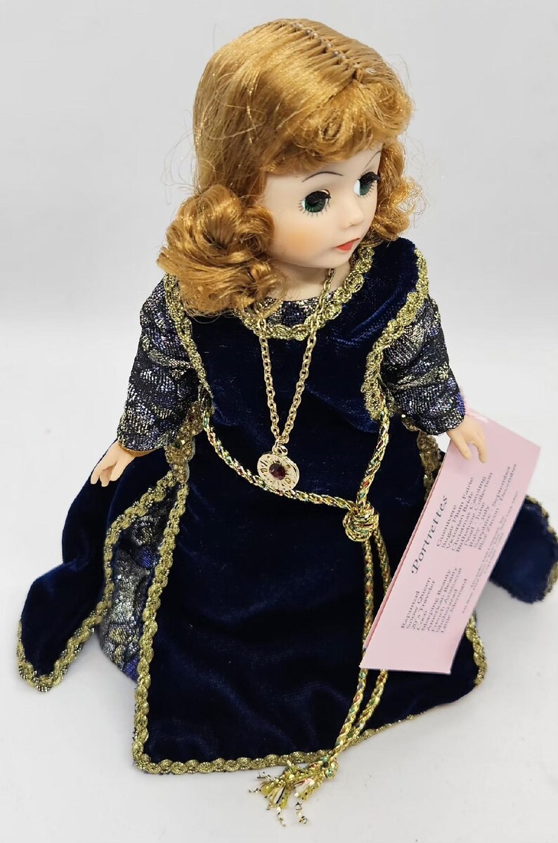 May include: A vintage doll with auburn hair and green eyes, wearing a dark blue velvet dress with gold trim. The doll has a gold necklace with a pendant and is holding a pink tag that reads "Portrettes".