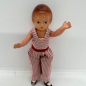 May include: A vintage doll with brown hair, blue eyes, and a red and white striped outfit. The doll is wearing black shoes.