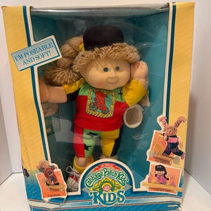 Puede incluir: Una muñeca Cabbage Patch Kids en una caja. La muñeca tiene cabello rubio, un sombrero negro y ropa colorida. La caja tiene texto que dice "I'M POSEABLE AND SOFT!" y "The Official Cabbage Patch Kids KIDS".