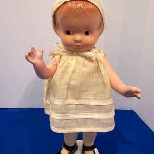 Può includere: Bambola vintage con viso dipinto, indossa un vestito color crema, un cappello e scarpe nere. La bambola ha un'espressione dolce ed è in posa con un braccio alzato. La bambola è su una base bianca.