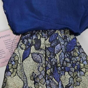 May include: A doll's outfit featuring a navy blue top and a skirt with a floral pattern in silver, blue, and gold. The skirt has a metallic sheen. A tag with text is partially visible.