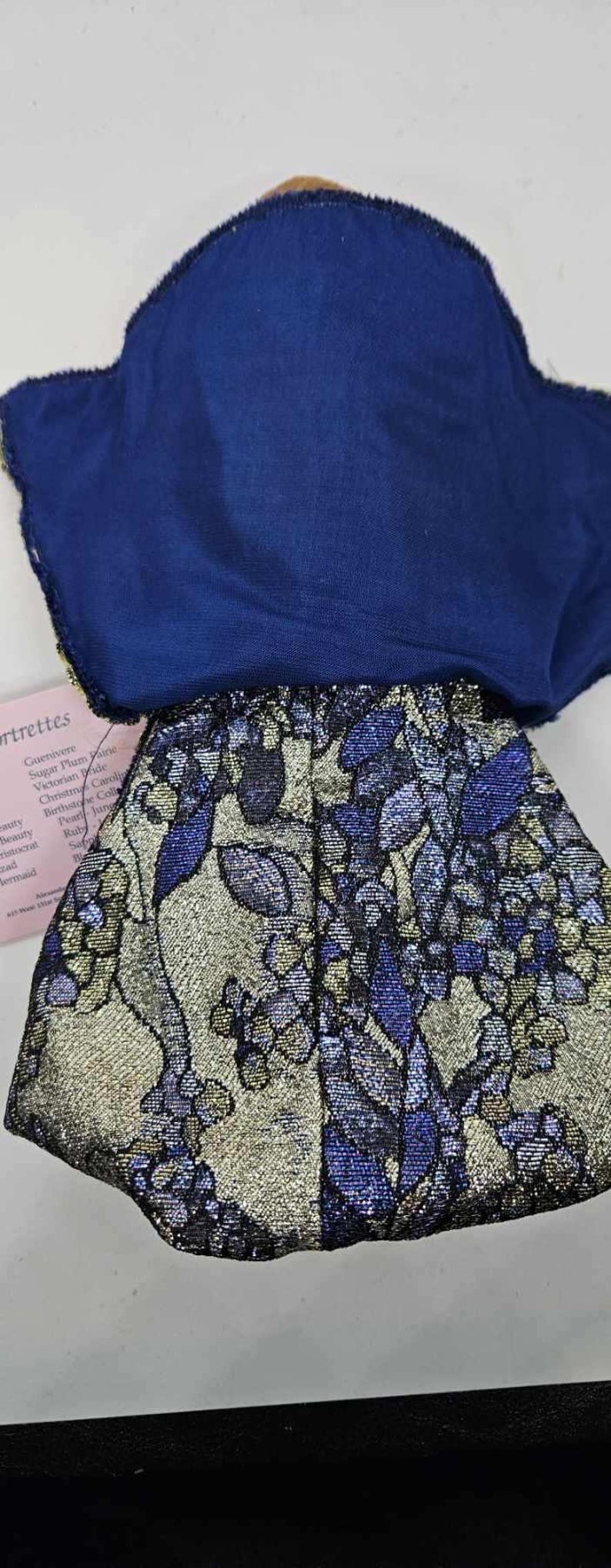 May include: A doll's outfit featuring a navy blue top and a skirt with a floral pattern in silver, blue, and gold. The skirt has a metallic sheen. A tag with text is partially visible.
