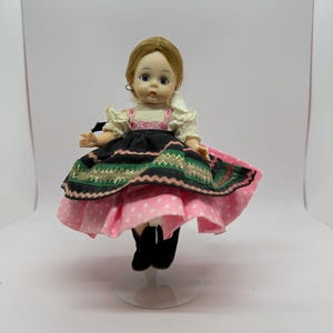 May include: A vintage doll with a painted face, wearing a traditional folk costume. The doll's dress features a black bodice, a pink polka-dot skirt, and a green and gold embroidered trim. The doll is approximately 6 inches tall.