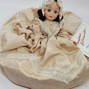 May include: A vintage doll with a cream-colored dress and matching bonnet. The doll has dark hair, blue eyes, and a delicate face. The dress is trimmed with lace, and the doll is holding a tag that reads "Madame Alexander".