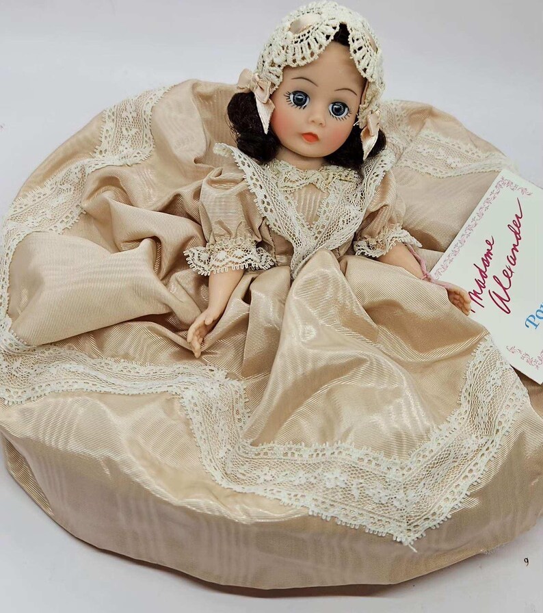 May include: A vintage doll with a cream-colored dress and matching bonnet. The doll has dark hair, blue eyes, and a delicate face. The dress is trimmed with lace, and the doll is holding a tag that reads "Madame Alexander".