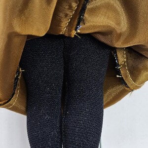 May include: Close-up of a doll's legs in black tights, with a gold-colored fabric above. The fabric is gathered and stitched with black thread. The doll's legs are straight and the tights are opaque.