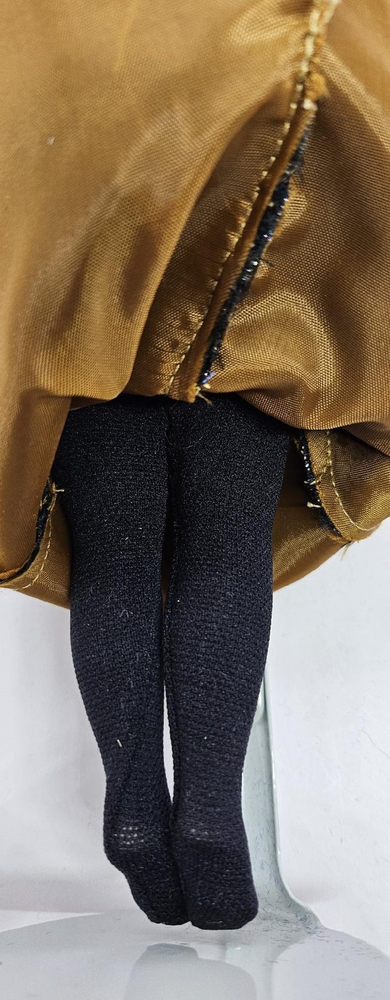 May include: Close-up of a doll's legs in black tights, with a gold-colored fabric above. The fabric is gathered and stitched with black thread. The doll's legs are straight and the tights are opaque.
