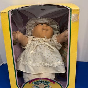 May include: A 1985 Cabbage Patch Kids Preemie doll with a round face and green eyes, wearing a cream-colored dress with a small floral pattern, a matching bonnet, and white pants, packaged in a yellow cardboard box with clear plastic window.