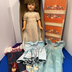 Könnte beinhalten: Eine Vintage-Puppe mit braunen Haaren und einem weißen Kleid, in ihrer Originalverpackung. Die Box ist orange mit dem Text "I BELONG TO". Weitere Artikel sind blaue Kleider, Jeans-Latzhosen und rote Schuhe.