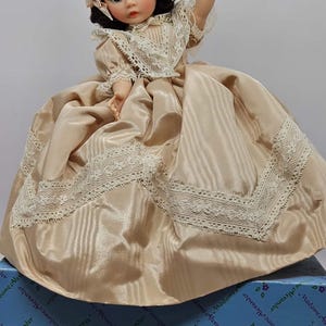 May include: A vintage Madame Alexander doll in a satin dress with lace trim and a matching bonnet. The doll has brown eyes and is holding a tag. The doll is on a blue box with the Madame Alexander logo.