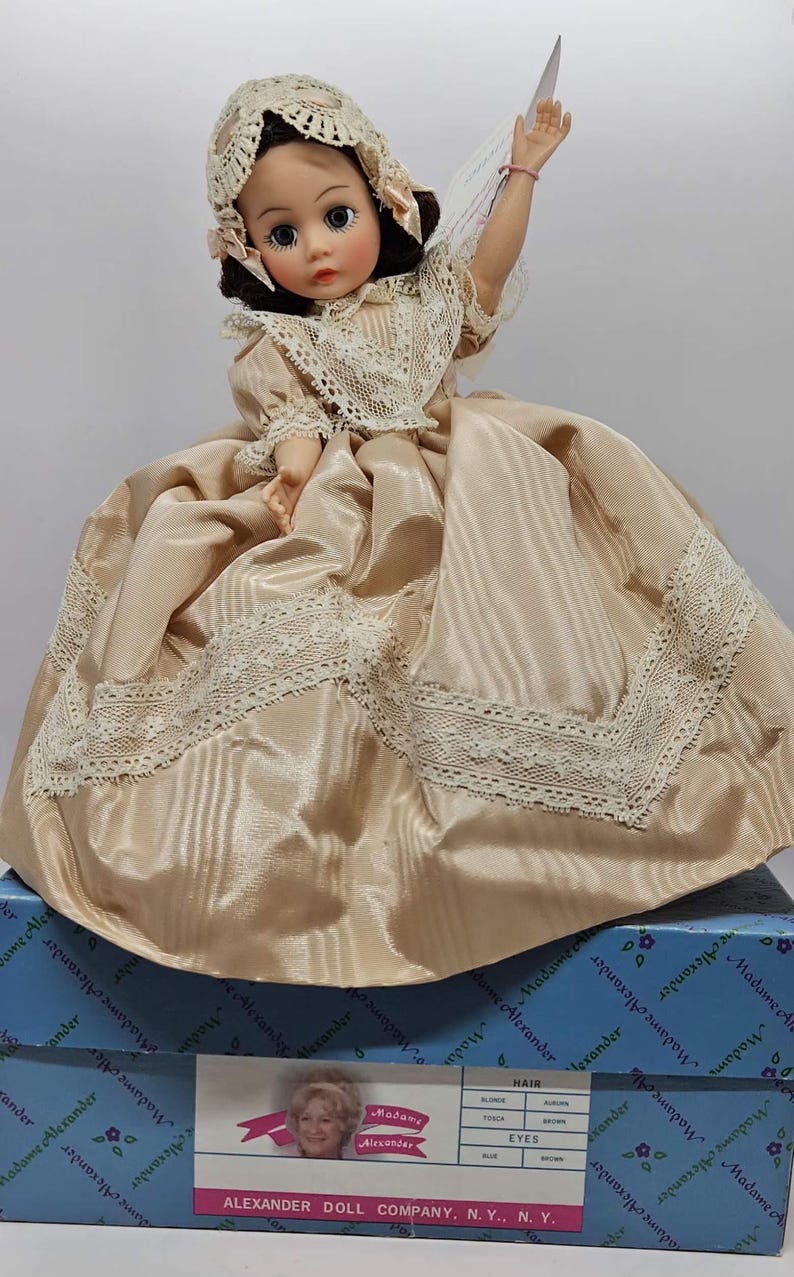 May include: A vintage Madame Alexander doll in a satin dress with lace trim and a matching bonnet. The doll has brown eyes and is holding a tag. The doll is on a blue box with the Madame Alexander logo.
