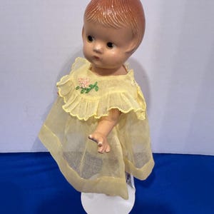 Può includere: Bambola vintage con viso dipinto e capelli castano-rossicci. La bambola indossa un vestito giallo con colletto a balze e dettaglio ricamato floreale. La bambola è montata su un supporto bianco.