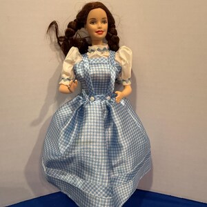 Könnte beinhalten: Eine Puppe mit braunem Haar, das zu zwei Zöpfen geflochten ist, trägt eine weiße Bluse mit kurzen Puffärmeln und ein blau-weiß kariertes Trägerkleid. Das Kleid hat eine hohe Taille mit zwei kleinen weißen Knöpfen am vorderen Oberteil.