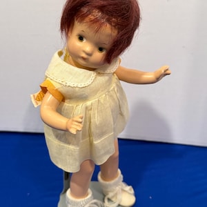 Könnte beinhalten: Eine Vintage-Puppe mit kastanienbraunem Haar und einem gelben Kleid. Die Puppe hat einen weißen Kragen und weiße Schuhe. Die Puppe steht auf einem grauen Ständer. Am Ärmel des Kleides der Puppe ist ein Etikett befestigt.