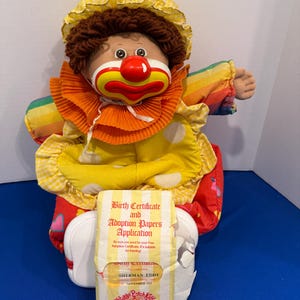 May include: A Cabbage Patch Kids Circus Kids doll with brown yarn hair, wearing a yellow gingham bonnet, a ruffled orange collar, and a yellow polka-dot dress with rainbow sleeves. It includes a paper Birth Certificate and Adoption Papers Application.