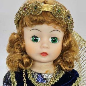 May include: A vintage doll with green eyes, red lips, and auburn hair styled in curls. The doll wears a gold-colored jeweled headband and a blue velvet dress with gold trim and a gold chain necklace. A decorative veil is visible.