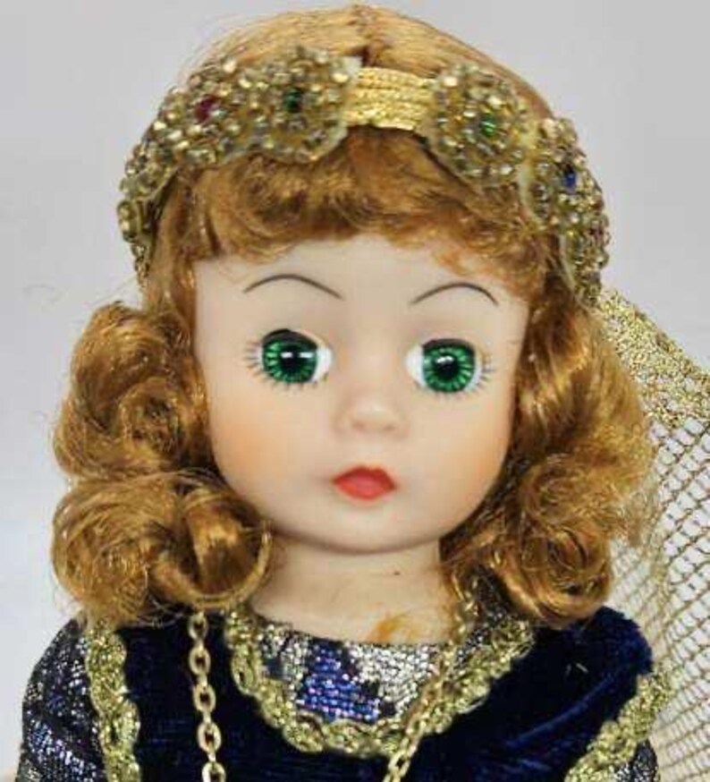May include: A vintage doll with green eyes, red lips, and auburn hair styled in curls. The doll wears a gold-colored jeweled headband and a blue velvet dress with gold trim and a gold chain necklace. A decorative veil is visible.