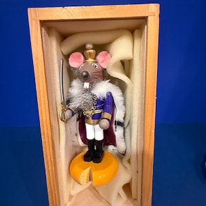 Könnte beinhalten: Ein hölzerner Nussknacker in Form eines Mäusekönigs mit goldener Krone, violetter Jacke und weißem, pelzbesetztem Umhang. Die Maus hält ein silbernes Schwert und steht auf einem gelben, käseförmigen Sockel in einer rechteckigen Holzkiste.