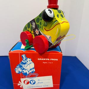 Könnte beinhalten: Vintage "Gran'pa Frog" Ziehspielzeug von Fisher-Price. Der Frosch hat einen grün-gelben Körper mit rotem Zylinder und roten Rädern. Das Spielzeug steht auf einer rot-blauen Box mit dem Namen und Logo des Spielzeugs. Für Kinder von 1 bis 4 Jahren.