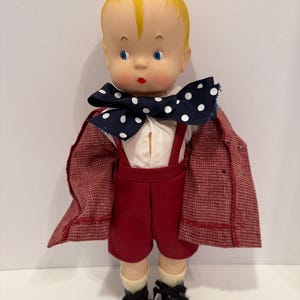 Può includere: Una bambola vintage con capelli gialli, occhi blu e labbra rosse. La bambola indossa una camicia bianca, pantaloncini rossi con bretelle, un papillon blu navy a pois bianchi, un blazer rosso, scarpe nere e calzini bianchi.