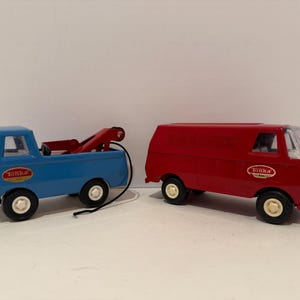 May include: Two vintage toy trucks: a blue tow truck with a red crane and a red van. Both have the "Tonka" logo. The tow truck has a black cable. The van has a clear windshield and windows. Both have black tires.