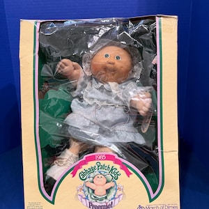 Könnte beinhalten: Eine Cabbage Patch Kids Preemie Puppe aus dem Jahr 1985 in Originalverpackung. Die Puppe hat ein rundes Gesicht, grüne Augen und trägt ein hellblaues Kleid mit weißen Punkten, eine Spitzenhaube und weiße Schuhe. Die Box zeigt das March of Dimes Logo.