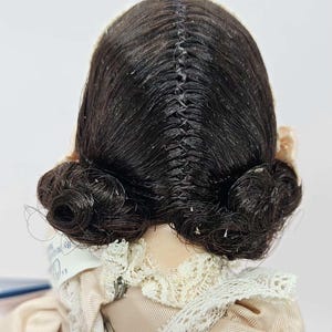 May include: A vintage doll with dark brown hair styled in two low buns. The hair is parted down the middle and sewn together. The doll is wearing a light pink dress with a lace collar.