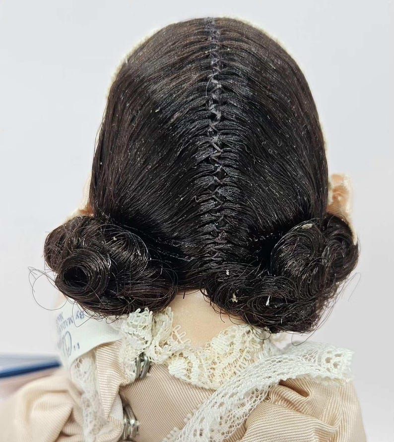 May include: A vintage doll with dark brown hair styled in two low buns. The hair is parted down the middle and sewn together. The doll is wearing a light pink dress with a lace collar.