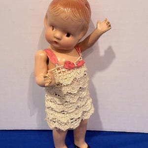 Può includere: Una bambola vintage con viso dipinto, capelli rossi e un vestito di pizzo color crema. La bambola ha scarpe nere e spalline rosa con un fiocco. La bambola è in piedi con un braccio alzato.