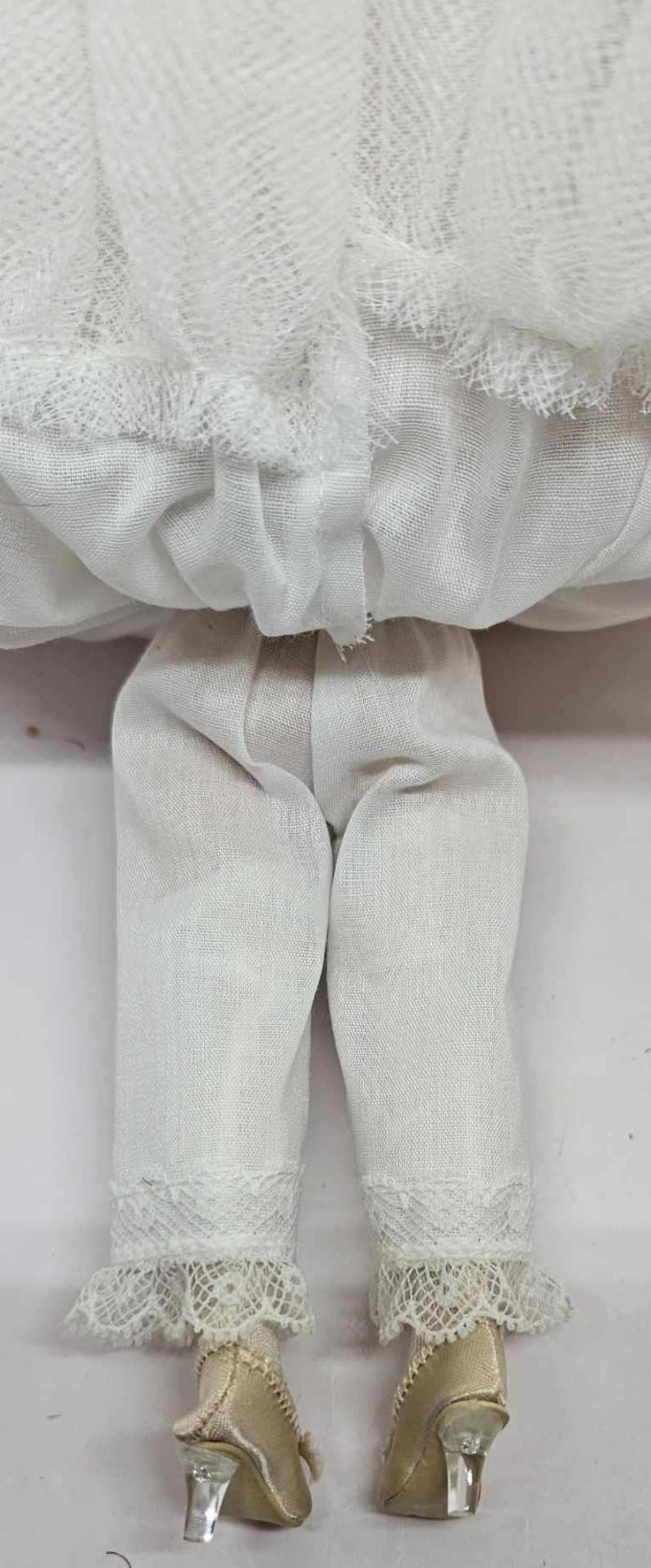 May include: A vintage doll's lower body, featuring white linen pants with lace trim at the ankles. The doll's legs end in gold-colored shoes with clear heels. The doll's skirt is made of a sheer, white fabric.
