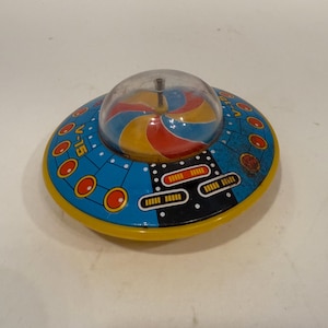 Może przedstawiać: Zabytkowy blaszany latający spodek w kolorze niebieskim, żółtym i czerwonym. Zabawka ma przezroczystą plastikową kopułę z obracającym się, kolorowym wzorem w środku. Na boku widnieje napis "V-15".