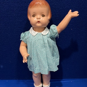 May include: A vintage doll with red hair, brown eyes, and a light blue dress with white floral print. The doll is wearing white socks and light blue shoes.