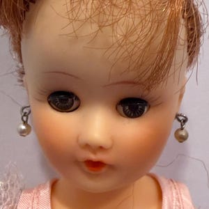 May include: Close-up of a vintage doll's face. The doll has red hair, dark eyes, and is wearing pearl earrings. The doll's lips are painted red, and it is wearing a pink dress.